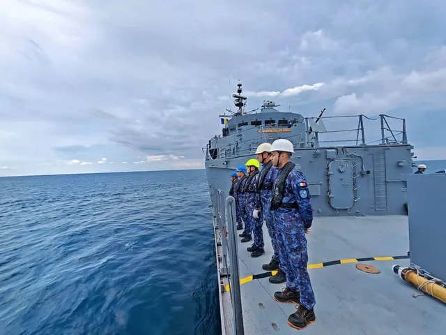 Câțiva militari în uniforme de camuflaj albastru stau pe puntea unui vas gri aflat pe o mare cu valuri mici sub un cer înnorat.