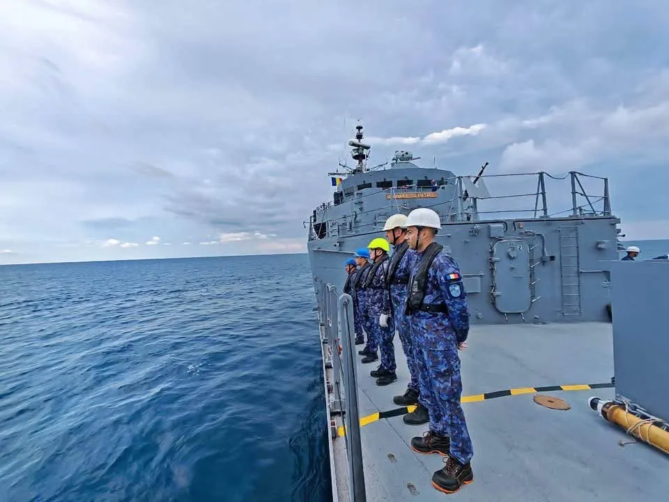 Câțiva militari în uniforme de camuflaj albastru stau pe puntea unui vas gri aflat pe o mare cu valuri mici sub un cer înnorat.