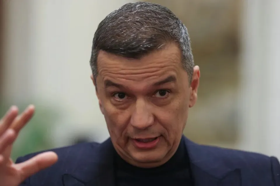Un bărbat cu părul scurt și gri poartă un sacou închis peste un tricou negru, având mâna stângă ridicată în prim-plan.