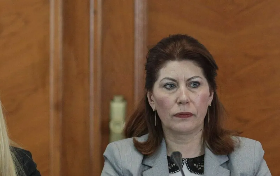 O femeie cu părul castaniu și o jachetă gri stă în fața unui fundal din panouri de lemn.