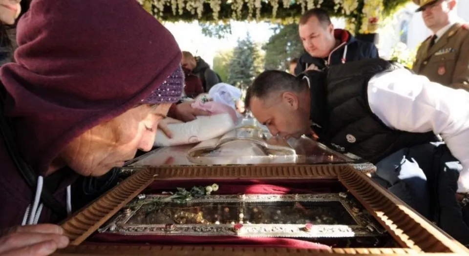 O femeie cu batic maro și un bărbat se apleacă pentru a săruta un obiect metalic decorat aflat într-o casetă de lemn sub un acoperiș.
