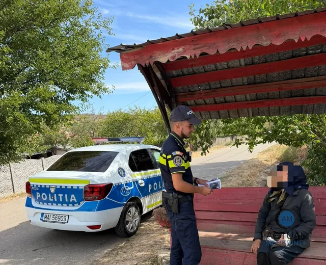 Un polițist în uniformă stă lângă o mașină de serviciu albastră și oferă un document unei persoane care stă pe o bancă.