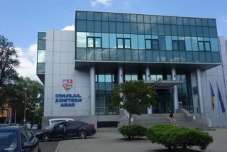 O clădire modernă cu fațadă de sticlă și panouri gri prezintă o intrare cu scări și câteva mașini parcate în prim-plan.