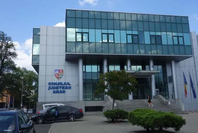 O clădire modernă cu fațadă de sticlă și panouri gri prezintă o intrare cu scări și câteva mașini parcate în prim-plan.