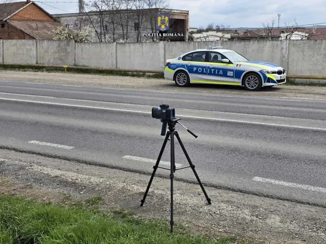 Un aparat foto montat pe un trepied negru este poziționat pe marginea unui drum asfaltat, în fața unei mașini de poliție albă, albastră și galbenă.