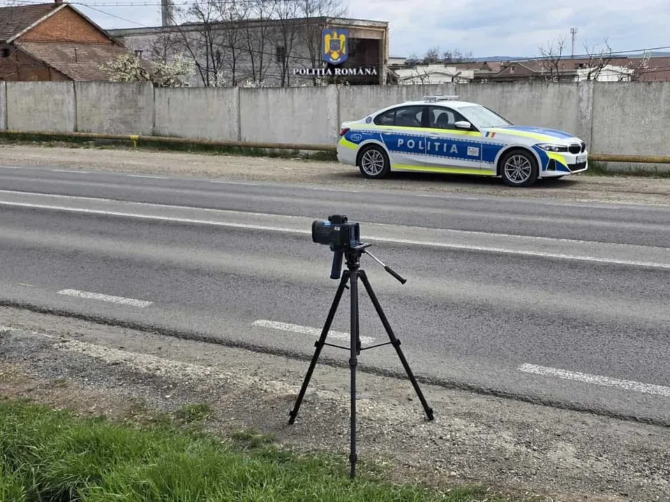 Un aparat foto montat pe un trepied negru este poziționat pe marginea unui drum asfaltat, în fața unei mașini de poliție albă, albastră și galbenă.