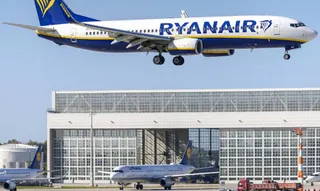 Un avion alb și albastru cu inscripția Ryanair zboară deasupra unui hangar mare și a unui avion aflat pe pistă.