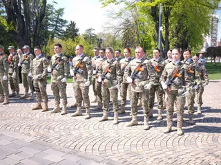 Un grup de militari în uniformă de camuflaj stă în rând pe o esplanadă pavată sub copaci verzi.