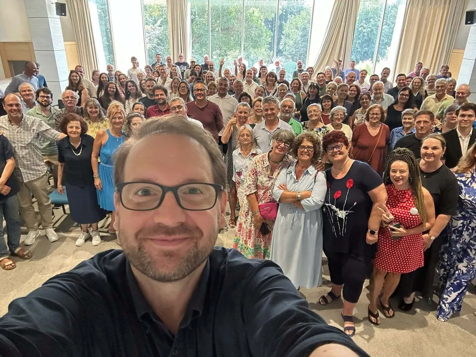 Un bărbat cu ochelari și cămașă închisă face un selfie în fața unui grup numeros de oameni care stau în interiorul unei clădiri luminoase.