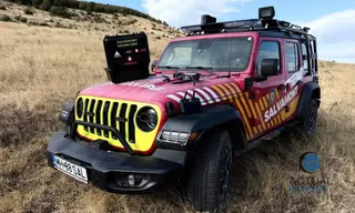 Un vehicul de teren roșu și galben cu inscripția Salvamont este parcat într-un câmp de iarbă uscată sub un cer înnorat.