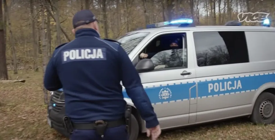 Un polițist cu un tricou albastru pe care scrie POLICJA stă lângă un vehicul alb cu lumini albastre aprinse și inscripția POLICJA pe lateral.