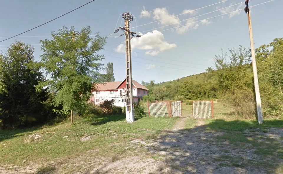O clădire cu acoperiș roșu și gard metalic se văd printre copaci, lângă o cale pietruită și stâlpi cu fire electrice.