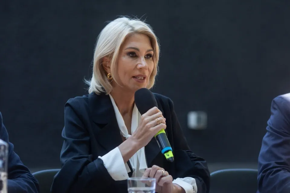O femeie cu părul blond, purtând o jachetă neagră și o cămașă albă, ține un microfon negru cu detalii verzi în mână.