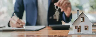 O persoană într-un costum albastru ține o cheie deasupra unei mașini de calcul, lângă o machetă de casă pe o masă de lemn.