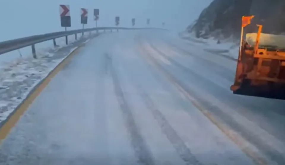 Un drum acoperit de zăpadă și gheață prezintă urme de roți și este delimitat de un parapet și semne de viraj pe partea stângă.