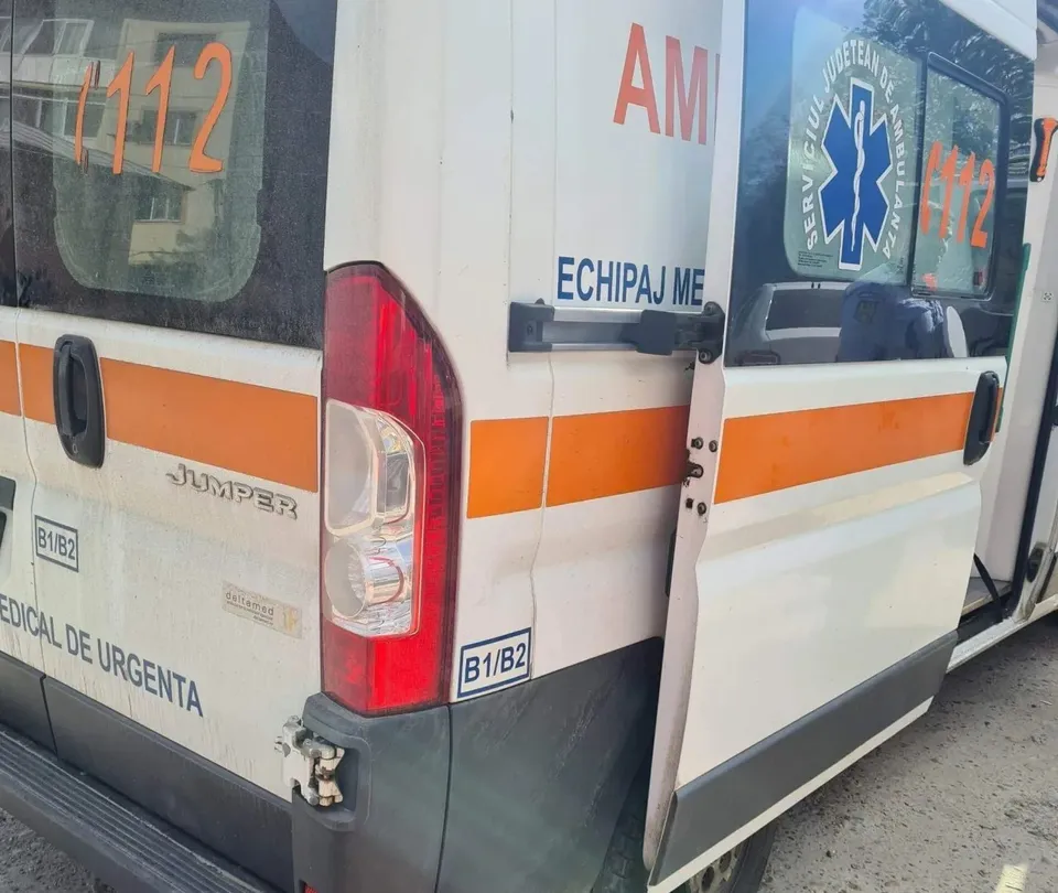 Partea din spate a unui vehicul alb cu benzi portocalii și inscripții medicale este vizibilă pe un fundal de asfalt.