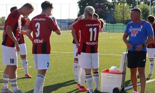 Câțiva jucători de fotbal în echipamente roșii și albe stau pe un teren de fotbal lângă un bărbat într-un tricou albastru și o ladă frigorifică.