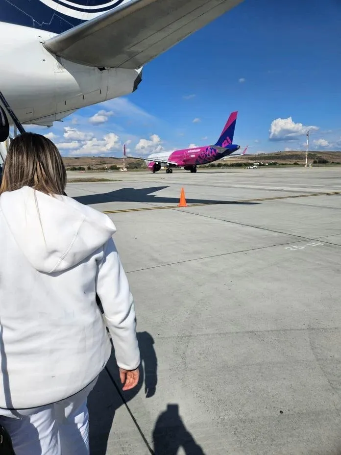 O persoană cu părul lung și haine albe privește un avion roz și alb parcat pe o pistă de beton sub un cer albastru.