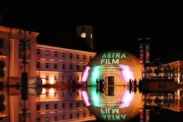 O sferă iluminată cu textul "ASTRA FILM" este reflectată într-o suprafață de apă lângă clădiri iluminate noaptea.