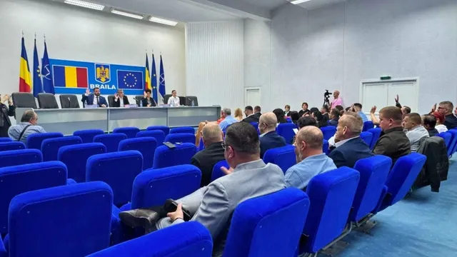 O sală cu rânduri de scaune albastre, audiență așezată și oficiali la o masă lungă, în fața unor steaguri și panouri.