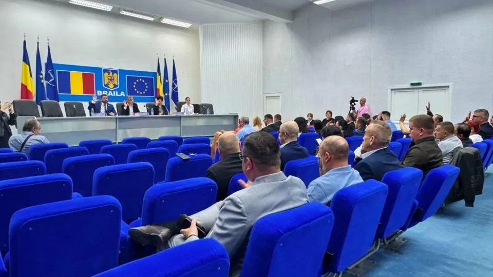 O sală cu rânduri de scaune albastre, audiență așezată și oficiali la o masă lungă, în fața unor steaguri și panouri.