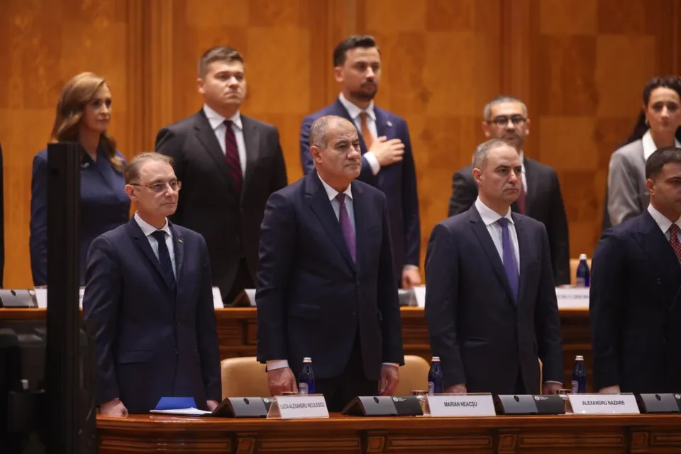 Un grup de bărbați și o femeie în costume oficiale stau în picioare în fața unor mese cu plăcuțe de identificare într-o încăpere cu panouri de lemn.