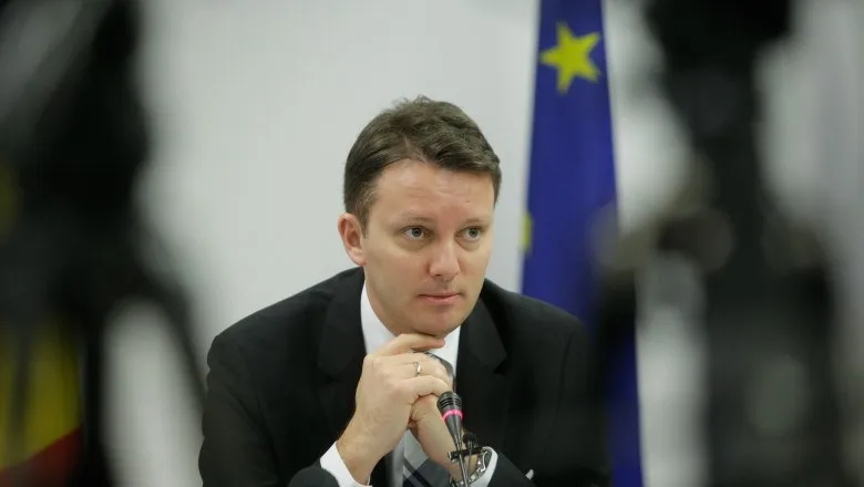Un bărbat cu părul scurt, îmbrăcat în costum negru și cămașă albă, stă cu mâinile sub bărbie în fața unui microfon.