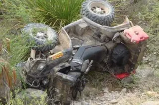 Un vehicul cu patru roți de culoare închisă este răsturnat pe o panta de teren accidentat, înconjurat de vegetație și pietriș.