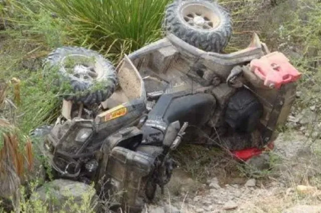 Un vehicul cu patru roți de culoare închisă este răsturnat pe o panta de teren accidentat, înconjurat de vegetație și pietriș.
