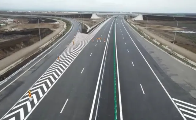 O porțiune de autostradă cu mai multe benzi de rulare, marcaje albe și verzi, este înconjurată de terenuri de construcție și pământ întins.