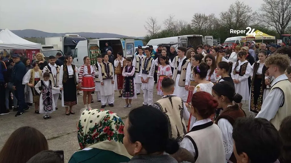 Un grup de persoane, inclusiv copii, poartă costume populare tradiționale și stau adunate pe un teren deschis în fața unor camioane albe.