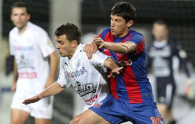 Un jucător de fotbal în echipament alb se confruntă cu un jucător în echipament albastru și roșu pe un teren de sport.