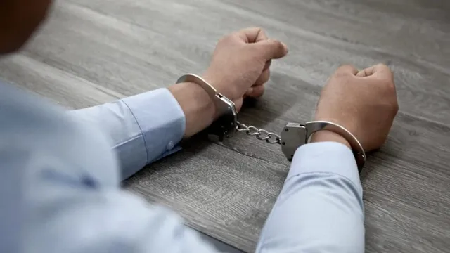 Un bărbat cu mâinile în spate este legat cu cătușe metalice deasupra unei suprafețe de lemn.