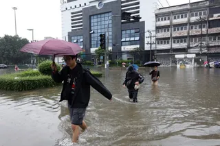 O persoană cu o umbrelă roz merge prin apă inundată pe un drum stradal lângă clădiri și vegetație.