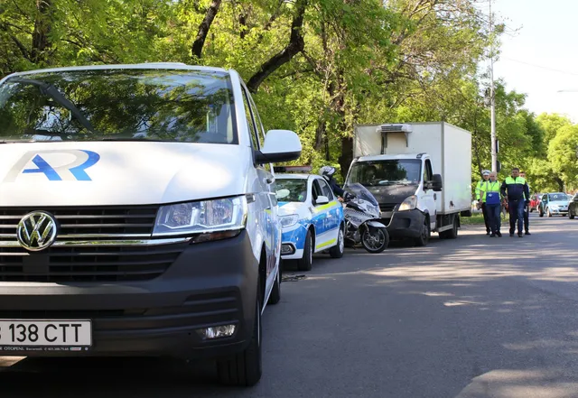 Un van alb, o mașină de poliție albastră și o motocicletă sunt parcate pe un drum lângă un camion alb și un grup de persoane.