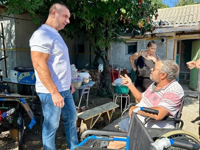 Un bărbat în tricou alb stă lângă un alt bărbat așezat într-un scaun cu rotile, în timp ce o femeie stă în fundal.