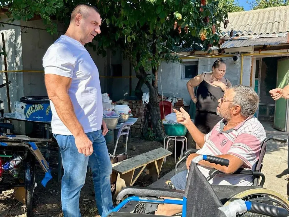Un bărbat în tricou alb stă lângă un alt bărbat așezat într-un scaun cu rotile, în timp ce o femeie stă în fundal.