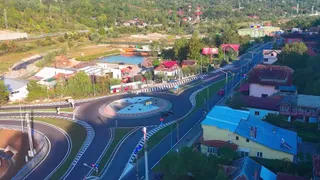 O vedere aeriană prezintă o intersecție de drum cu un sens giratoriu, asfalt gri, linii albe marcate și case cu acoperișuri colorate în apropiere.