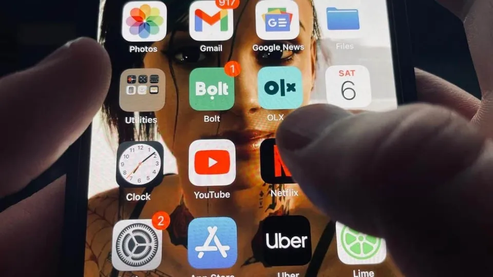 O persoană ține un telefon mobil cu ecranul pornit, afișând diverse aplicații, inclusiv Gmail, Google News, Bolt și OLX. Pe fundalul ecranului se vede o imagine cu o femeie.