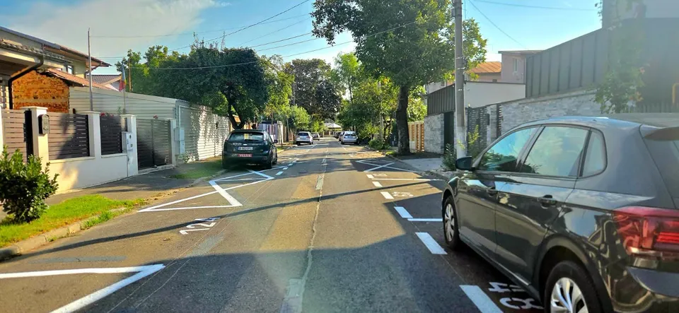 O stradă asfaltată cu linii albe marcate pe carosabil, având mașini parcate pe margini și case cu garduri în fundal.