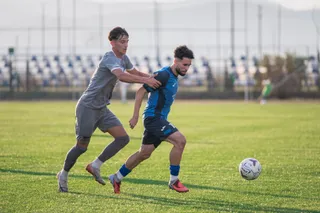 Un jucător într-un echipament albastru conduce mingea în fața unui adversar într-un echipament gri pe un teren de fotbal cu iarbă verde.