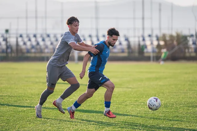 Un jucător într-un echipament albastru conduce mingea în fața unui adversar într-un echipament gri pe un teren de fotbal cu iarbă verde.