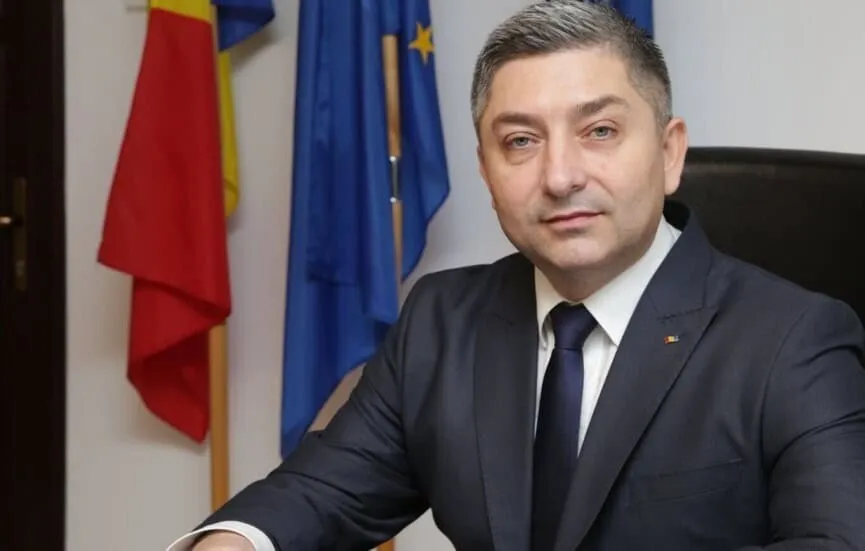Un bărbat cu părul scurt și gri, purtând un costum închis la culoare și cravată, stă în fața unor steaguri colorate.