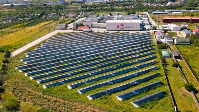 O vedere aeriană prezintă rânduri lungi de panouri solare instalate pe un teren verde lângă diverse clădiri industriale.