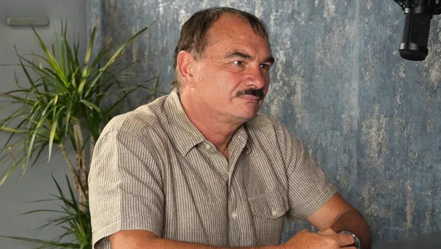Un bărbat cu mustață și cămașă cu mânecă scurtă stă așezat în fața unui fundal texturat și a unei plante verzi.