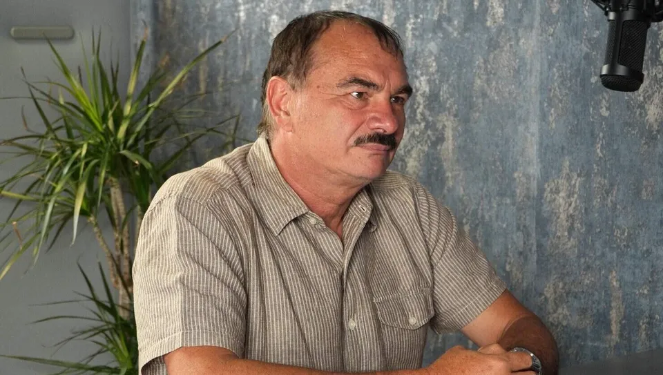 Un bărbat cu mustață și cămașă cu mânecă scurtă stă așezat în fața unui fundal texturat și a unei plante verzi.