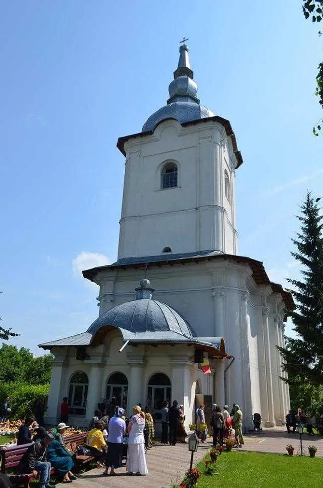 O clădire albă cu un turn înalt și un cupolă se ridică sub un cer senin, în fața căreia stau grupuri de oameni pe un teren verde.