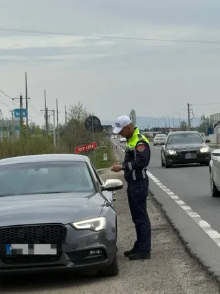 Un polițist în uniformă stă pe marginea drumului lângă o mașină gri, privind spre interiorul vehiculului.