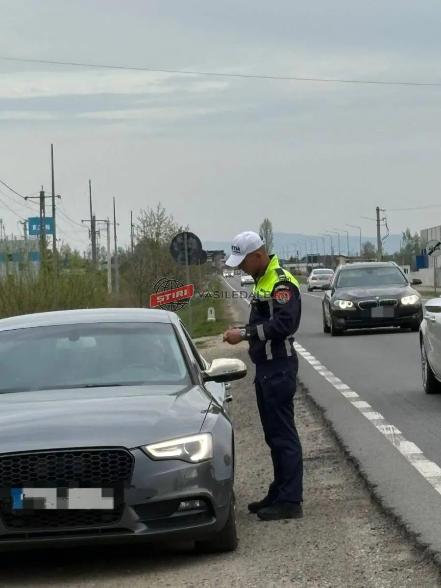 Un polițist în uniformă stă pe marginea drumului lângă o mașină gri, privind spre interiorul vehiculului.