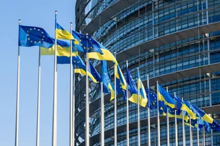 Mai multe steaguri ale Uniunii Europene și ale Ucrainei sunt arborate pe catargi în fața unei clădiri moderne cu fațadă de sticlă.